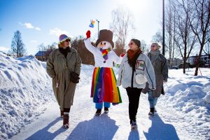 Arctic pride maskotti kävelemässä sateenkaarilippu kädessä kolmen henkilön kanssa talvisessa maisemassa